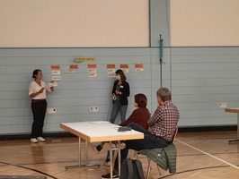 In der Turnhalle präsentieren zwei Frauen das Projekt anhand von kleinen Zetteln, die sie an die Wand pinnen. Im Vordergrund sitzen zwei Personen, die gespannt auf die Wand blicken.